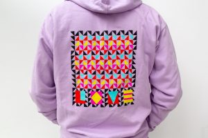 Close up of the back of a man wearing a hooded sweatshirt printed with a Jeffrey Gibson-design colorful triangle patterned graphic spelling LOVE.